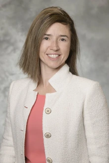 Portrait of a fair-skinned woman with soulder-length brown hair smiling. She is wearing a white blaser.