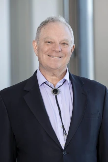 Portrait of Dr. Geoffrey Rubin smiling and wearing a suit coat, collared shirt and bolo tie.