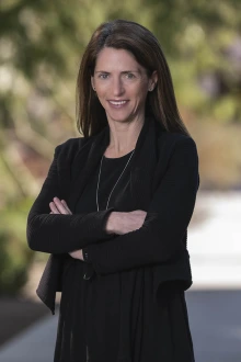 White woman in dark blouse with arms folded in outdoor setting. 