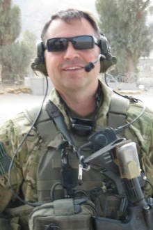 A man in full Army tactical gear and wearing dark glasses and a radio headset stands in an outside setting. 