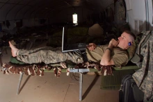 A male soldier lies on a cot while watching a laptop computer and wearing headphones. 