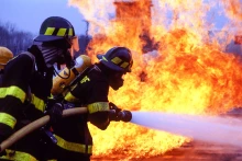 Two firefighters wearing protective gear and oxygen tanks spray on blaze