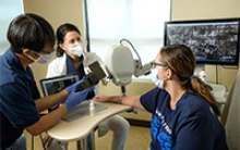 Drs. Clara Curiel Lewandrowski and Dongkyun Kang prepare to do a side-by-side test of the confocal microscope and the portable unit to compare the image quality. Dr. Curiel is aligning the larger device, while Dr. Kang prepares to aim the handheld device at a nearby spot on Savannah Barber’s arm. Barber is a medical assistant at Banner – University Medical Center Tucson. (Photo: Kris Hanning/University of Arizona Health Sciences)