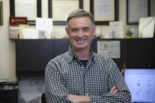 A man standing in an office with computer and awards in the background