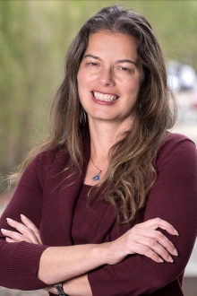 Portrait of a woman with long brown hair standing outside smiling with arms folded.